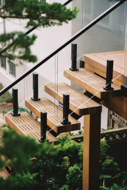 Slatted wood treads with black glass posts turn at a landing among evergreen foliage.