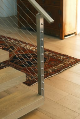 Brushed stainless post with cable infill at the foot of light oak floating treads on a rug.