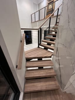 Wood floating treads turn on a black mono stringer with cable railing beside a marble wall.