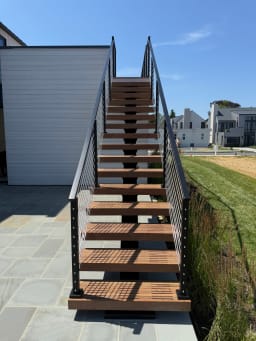 Straight-on view of slatted hardwood floating treads on a mono stringer rising between black cable railings from a bluestone patio.