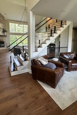 Wood stair with white risers and frameless glass on black talons rising beside a leather-furnished family room.