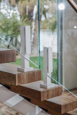 Bright white ascend talons clamp frameless glass to thick wood treads beside a window.