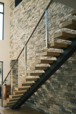 Light oak treads on a black mono stringer with stainless cable railing climb a stacked-stone wall.