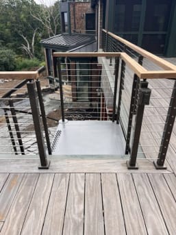Black cable railing with wood cap frames a stair opening on a weathered wood deck.