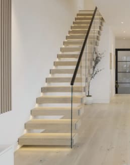 Light oak cantilever treads with under-tread LED lighting and a glass railing with black handrail rise along a white wall.