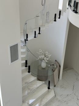 Curving white marble stair with curved frameless glass on black talons around a foyer table.