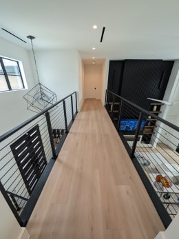 Upper floor oak bridge walkway lined on both sides by black cable railing above an open living area.