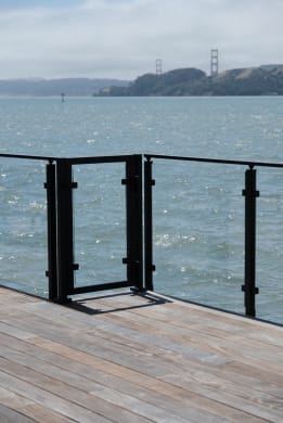Black post-to-post glass railing and matching gate on a wood dock deck overlooking the bay and a distant bridge.