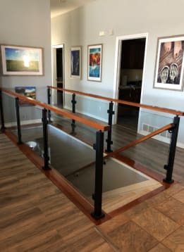 Black posts with glass panels and a stained wood top rail surround a stair opening in a hallway with framed art.