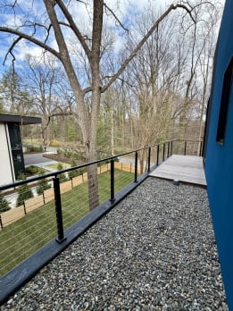 Black surface-mount cable railing lines a gravel and ipe walkway beside a modern blue home