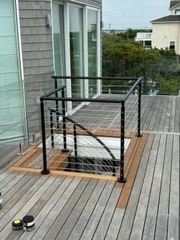 Black cable railing guards a rooftop deck hatch with a curving stair below on a coastal home