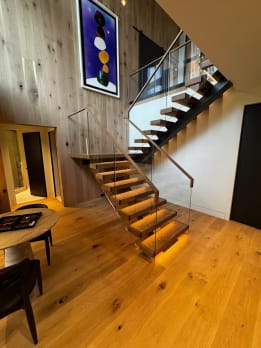 Thick oak treads with warm under-tread LED lighting float on black stringers beside frameless glass with a wood handrail in a double-height wood-paneled room.