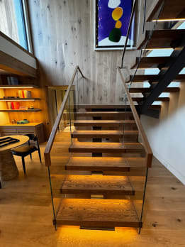 A straight run of oak floating treads with LED under-lighting rises between frameless glass guards topped with wood handrails.