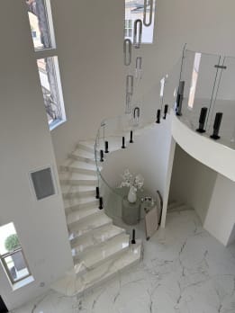 Curved glass railing on black talon clamps follows a white marble spiral stair under a crystal pendant.