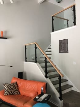 Black posts with glass panels and wood handrail guard a gray-tread stair and upper landing.