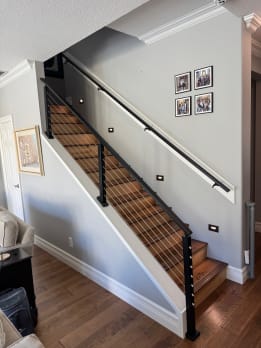 A black wall-mounted metal handrail opposite black cable railing on a stained wood stair.