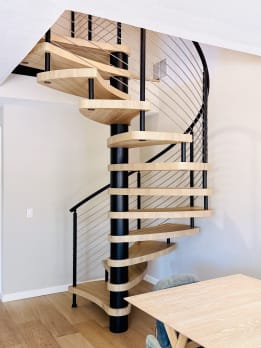 Close view of a spiral staircase with pale wood pie treads on a black center column and black curved cable railing.