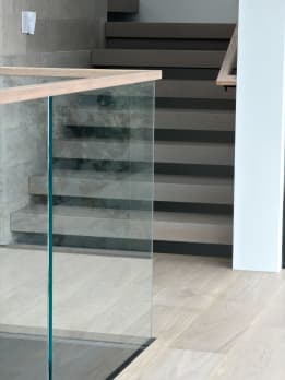 Gray-washed stack stair behind a frameless glass panel with a light wood cap rail.