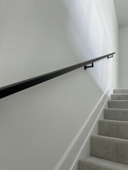 A slim black wall-mounted handrail runs along a white wall above a carpeted stair.