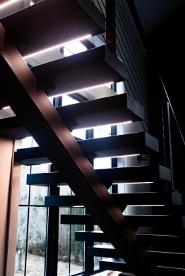 Looking up at dark floating treads with glowing LED strips on a dark mono stringer by tall windows.
