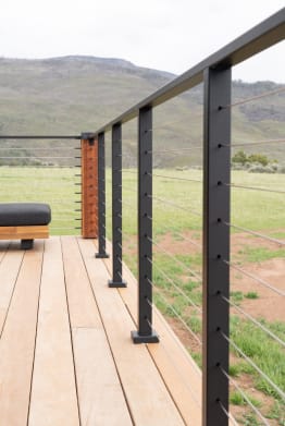 Black side-mounted posts with cable infill and black top rail along a light wood deck facing open hills.