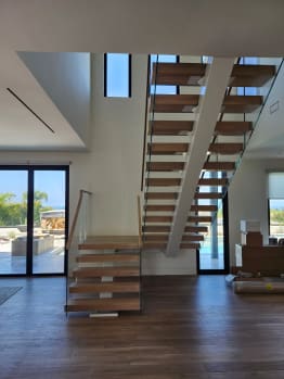 White oak floating stair with glass railings rising through a bright great room with ocean views