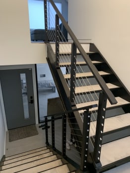 Black double posts with stainless cables and a black handrail start at the base of an interior steel-and-wood stair.