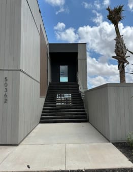 Wide black steel open-riser stair rises between gray-clad walls toward an upper entry under blue sky.