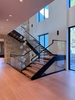 Light oak treads on black stringers with frameless glass railing and black handrail rise in a tall entry with a stone wall and orb chandelier.