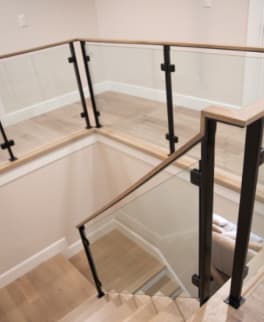 Black posts holding glass panels beneath a continuous light oak handrail wrapping a stair opening.