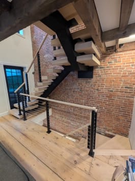 Pale oak floating stair on a black mono stringer with cable railing beside exposed brick and timber beams