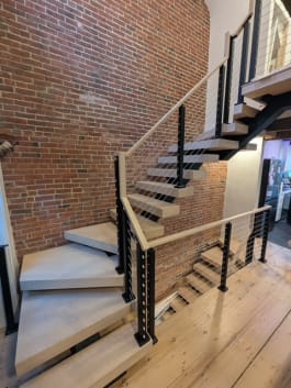 Switchback floating stair with pale oak treads and black cable railing along an exposed brick wall
