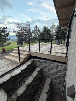 Black cable railing on a composite deck and steps above a stone window well, with mountain pasture beyond