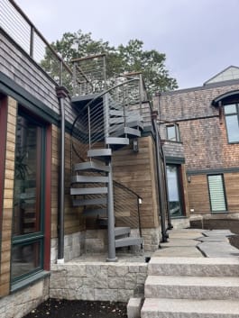 Gray steel spiral stair with cable railing climbs to a roof deck beside wood siding and granite steps.