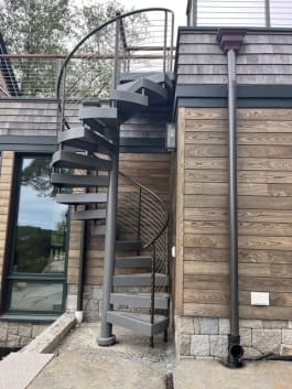 Gray steel spiral stair with curved cable railing beside cedar shingle and wood-clad walls.