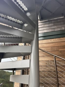 Underside of gray steel spiral treads and center column with cable railing against wood siding.