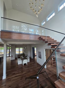Walnut floating treads with frameless glass railing and black handrail lead to an open balcony above a dining area.