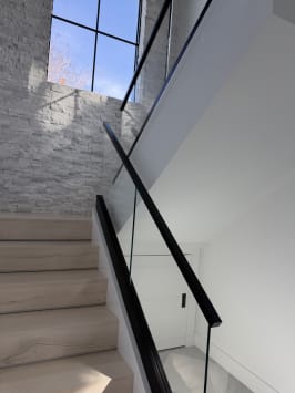 A white oak stair rises beside frameless glass on a kneewall with a black handrail toward a stone accent wall.