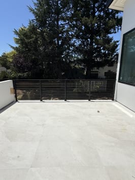 Black horizontal rod railing spans a pale stone terrace of a white modern home beneath tall trees.