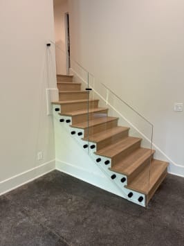 Frameless glass panels mounted with black standoff pins along a white oak closed stair in a bright stairwell.