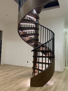 A dark oak wood-wrapped spiral stair with black baluster railing and LED tread lights in a white room.