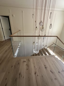 A frameless glass guard with an oak cap wraps a stair opening beneath a cascading pendant light in a paneled hallway.