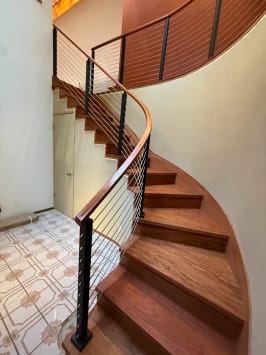 A curved wood stair rises with black-post cable railing and a bent wood handrail along a curved wall.