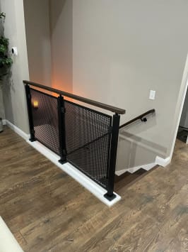 Black perforated metal panels with a dark wood cap guard a stair opening on a wood floor.