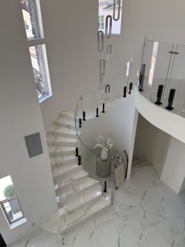Curved glass panels on black talon spigots follow a sweeping white marble stair beneath a crystal chandelier.