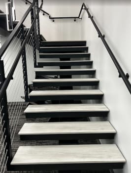 Gray treads on a black mono stringer between black cable railing and a wall-mounted black handrail in an office stairwell.