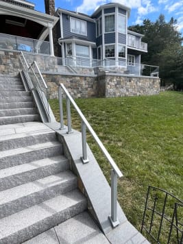 Granite steps with glass base rail railing rise toward a blue house, with an aluminum post railing in the foreground