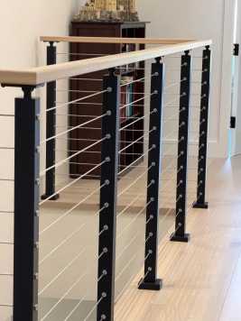 Close view of black cable railing posts and a white oak cap along a hallway