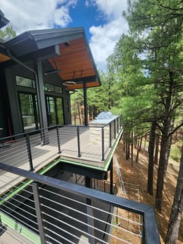 Black cable railing wraps a composite deck and stone-clad outdoor kitchen among tall pines