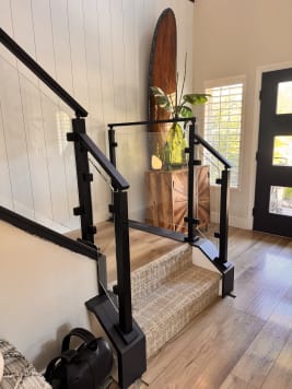 Black post glass railing climbs a carpeted stair from a wood-floored entry